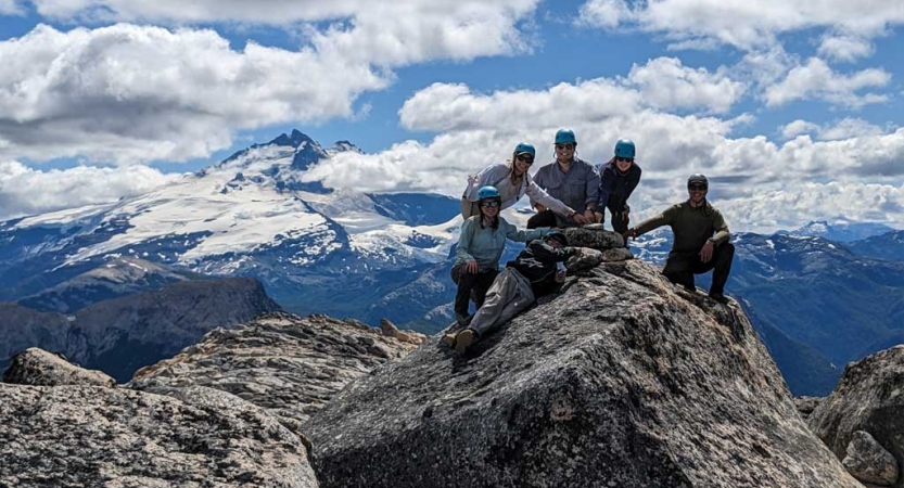 A group of people wearing helmets stand on a rocky peak. There is a vast mountain view in the background. 